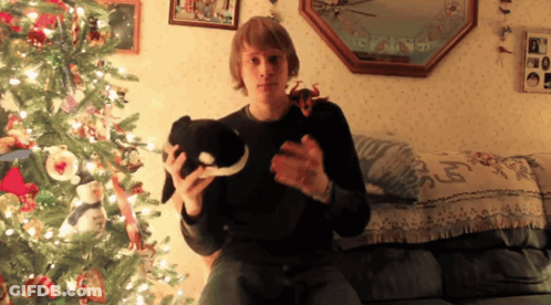 A Man Is Sitting On A Couch Holding A Stuffed Animal In Front Of A Christmas Tree GIF