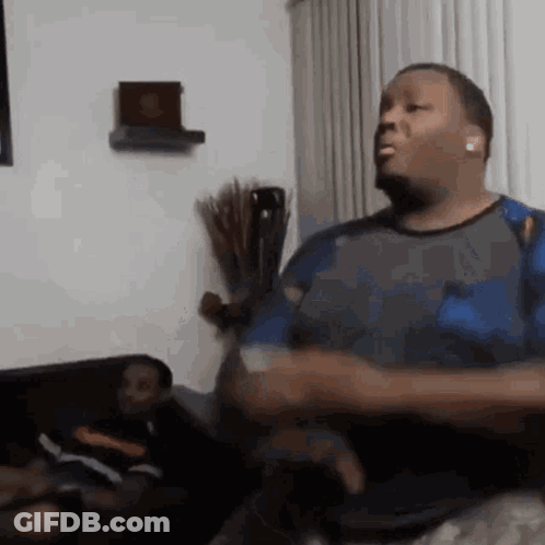 A Man Is Sitting On A Couch In A Living Room Playing A Video Game While Another Man Watches . GIF
