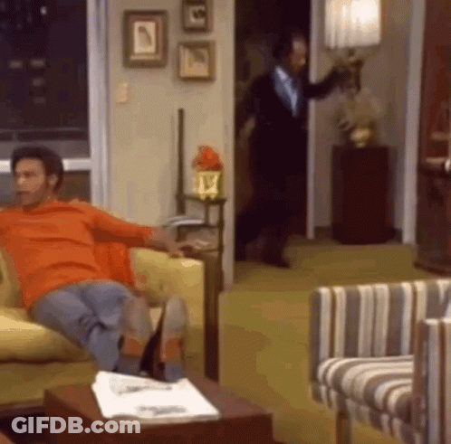 A Man Is Sitting On A Couch In A Living Room While A Man In A Suit Is Standing In The Doorway . GIF