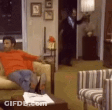 A Man Is Sitting On A Couch In A Living Room While A Man In A Suit Stands In The Doorway . GIF