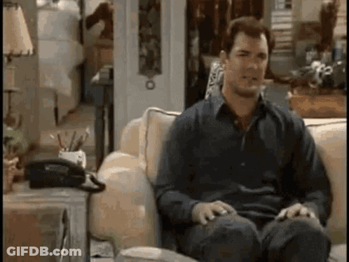 A Man Is Sitting On A Couch In A Living Room With A Telephone On The Table . GIF