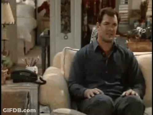 A Man Is Sitting On A Couch In A Living Room With His Legs Crossed And A Telephone On The Table . GIF