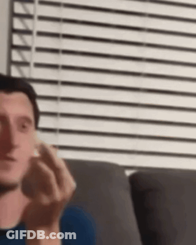 A Man Is Sitting On A Couch In Front Of A Window With Blinds And Holding Something In His Hand . GIF