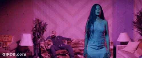 A Man Is Sitting On A Couch While A Woman In A Blue Dress Stands In Front Of Him . GIF