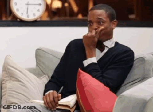 A Man Is Sitting On A Couch With A Book And A Clock Behind Him GIF