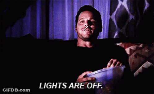 A Man Is Sitting On A Couch With A Bucket Of Popcorn And The Words Lights Are Off Above Him GIF