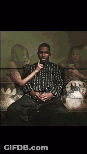 A Man Is Sitting On A Couch With A Woman Behind Him Holding His Hand To His Mouth GIF