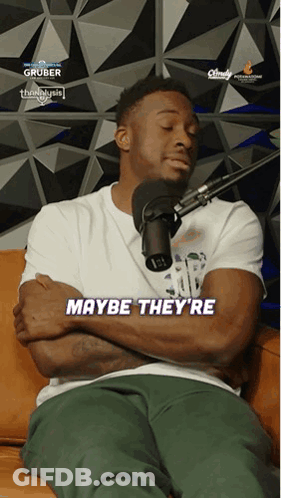 A Man Is Sitting On A Couch With His Arms Crossed And Says Maybe They 'Re In Front Of A Microphone GIF