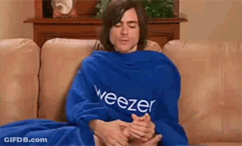 A Man Is Sitting On A Couch Wrapped In A Blue Blanket With The Word Weezer On It . GIF