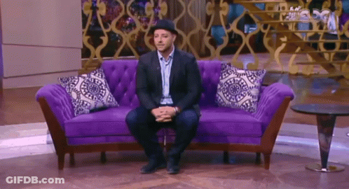 A Man Is Sitting On A Purple Couch With A Hat On GIF