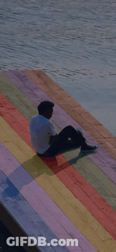 A Man Is Sitting On A Rainbow Colored Dock Overlooking A Body Of Water GIF