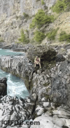 A Man Is Sitting On A Rock Near The Water . GIF