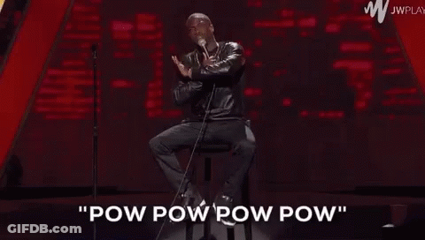 A Man Is Sitting On A Stool On A Stage Holding A Microphone And Talking Into It . GIF