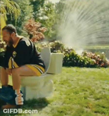 A Man Is Sitting On A Toilet In The Grass With A Sprinkler Spraying Water GIF