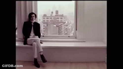 A Man Is Sitting On A Window Sill In Front Of A Window With A View Of A City . GIF