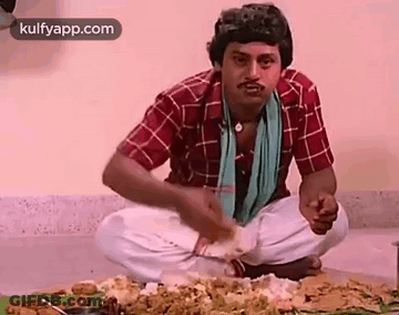 A Man Is Sitting On The Floor Eating Food From A Table . GIF