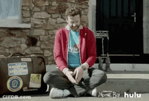 A Man Is Sitting On The Ground In Front Of A Stone Building . GIF