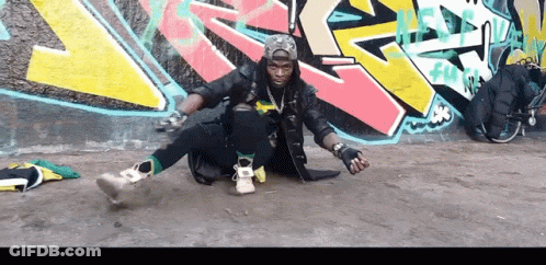 A Man Is Sitting On The Ground In Front Of A Wall With Graffiti On It That Says ' Fun ' GIF