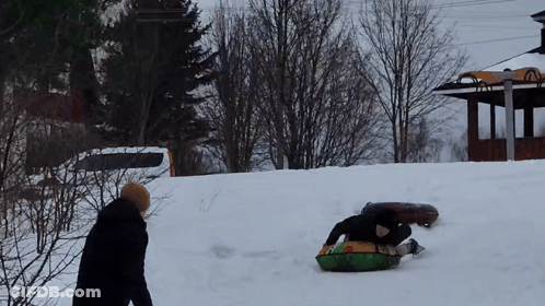 A Man Is Sledding Down A Snow Covered Hill While Another Man Watches GIF