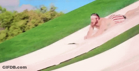 A Man Is Sliding Down A Water Slide In A Park . GIF