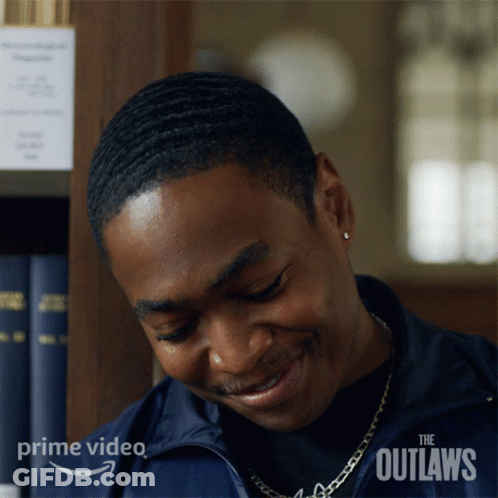A Man Is Smiling In Front Of A Bookshelf With The Outlaws Written On The Bottom Right Corner GIF