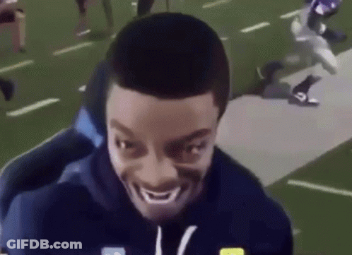 A Man Is Smiling On A Football Field While A Football Player Is Running Behind Him . GIF