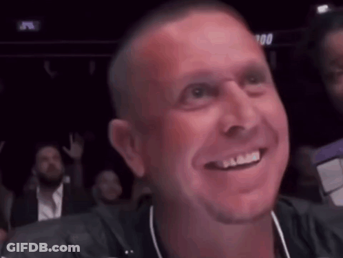 A Man Is Smiling While Sitting In A Crowd Of People . GIF