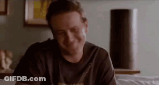 A Man Is Smiling While Sitting On A Bed In A Room . GIF