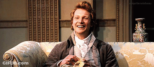 A Man Is Smiling While Sitting On A Couch With A Vase In The Background .. GIF