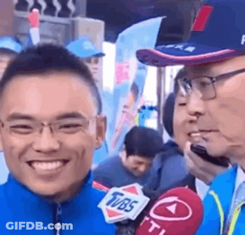 A Man Is Smiling While Talking Into A Microphone While Wearing Glasses And A Hat . GIF
