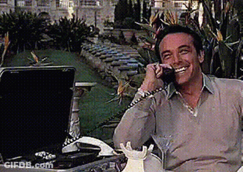 A Man Is Smiling While Talking On A Phone Next To A Briefcase GIF