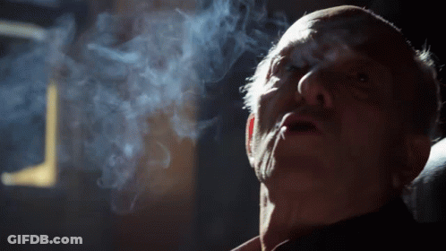 A Man Is Smoking A Cigarette In A Dark Room With Smoke Coming Out Of His Mouth GIF