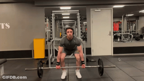 A Man Is Squatting Down Holding A Barbell In A Gym With The Word Ts On The Wall GIF