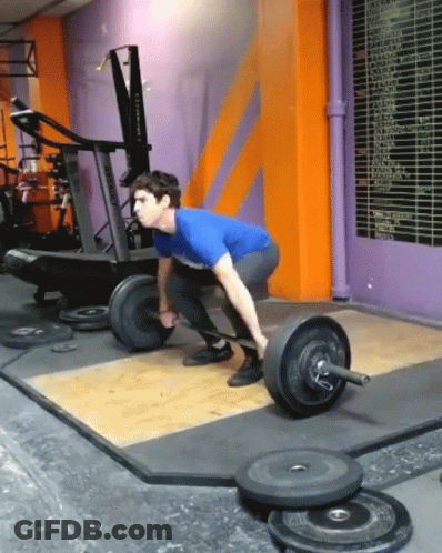 A Man Is Squatting Down To Lift A Barbell In A Gym With A Purple Wall Behind Him GIF