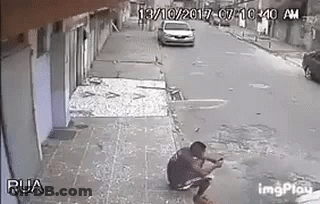 A Man Is Squatting On The Sidewalk While A Car Is Driving By . GIF