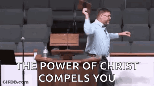 A Man Is Standing At A Podium In Front Of A Microphone Holding A Bible In His Hand . GIF