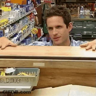 A Man Is Standing Behind A Counter In A Store Looking Over It . GIF