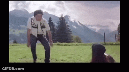 A Man Is Standing In A Field With Mountains In The Background While A Woman Looks On GIF