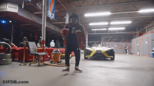 A Man Is Standing In A Garage With A Slingshot Car In The Background GIF