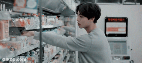 A Man Is Standing In A Grocery Store Reaching For A Bottle Of Water . GIF