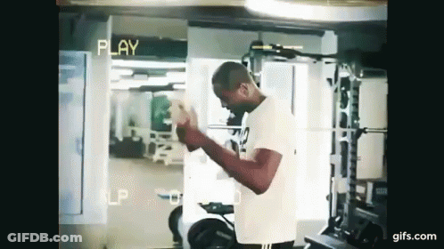 A Man Is Standing In A Gym Holding A Dumbbell In His Hands . GIF