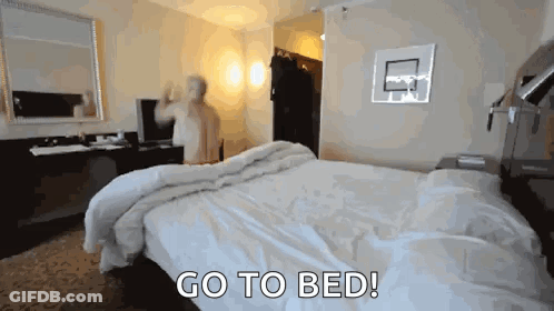 A Man Is Standing In A Hotel Room With The Words Go To Bed Behind Him GIF