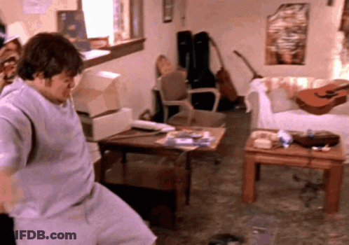 A Man Is Standing In A Living Room With A Guitar On A Table GIF