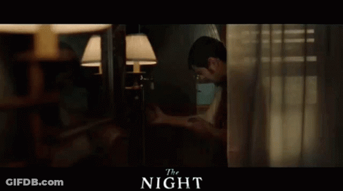 A Man Is Standing In A Room Behind A Curtain And Looking Out A Window . GIF