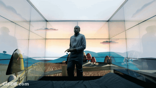 A Man Is Standing In A Room With A Painting Of A Landscape On The Wall GIF