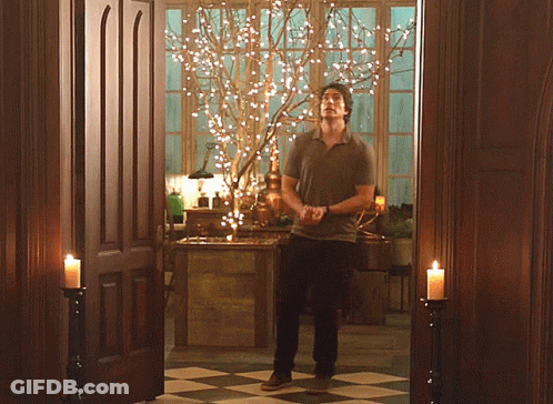 A Man Is Standing In A Room With Candles And A Tree With Lights On It GIF