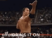 A Man Is Standing In A Wrestling Ring With His Arm In The Air And The Words `` Bring It On '' Behind Him . GIF