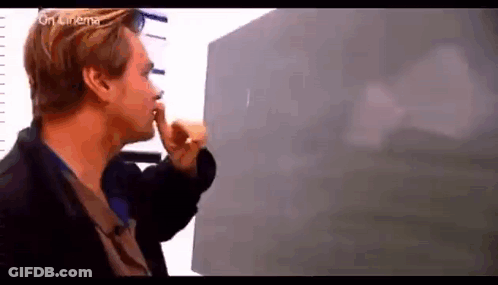 A Man Is Standing In Front Of A Blackboard Holding His Finger To His Lips . GIF
