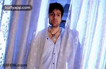 A Man Is Standing In Front Of A Blue Curtain And Looking At The Camera . GIF