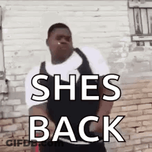 A Man Is Standing In Front Of A Brick Wall With The Words `` She 'S Back '' Written On His Chest . GIF